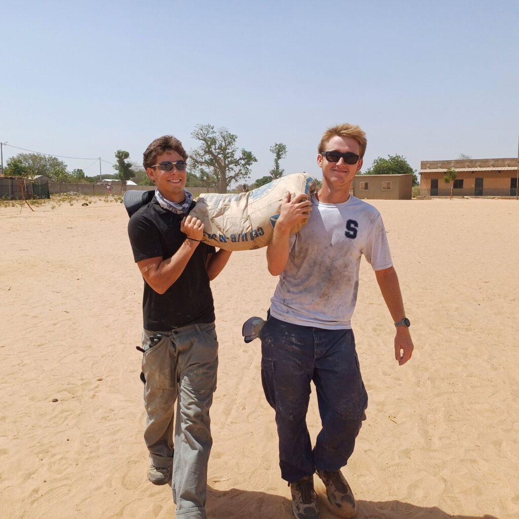 Greyson (Left) helps carry a bag of cement on Trek in Senegal.